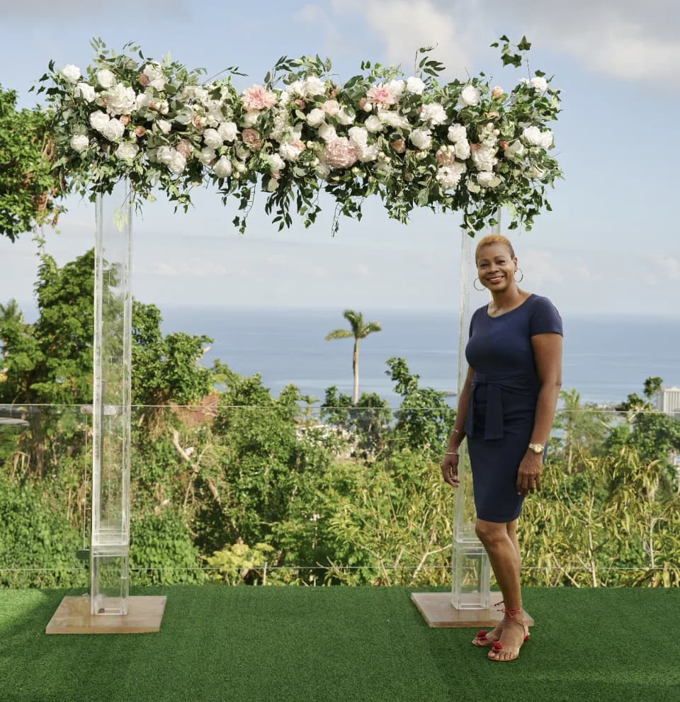 Michelle, host of Bay Vista Villa, beside a floral arch with the bay of Ocho Rios behind her